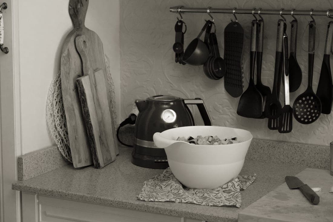 Wooden cutting boards of several sizes leaning against a kitchen backsplash beside a kettle and a mixing bowl